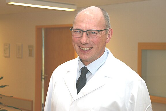 Ein lächelnder Mann mit Brille trägt weißen Kittel und Krawatte, steht in einem hellen Klinikflur und blickt in die Kamera.