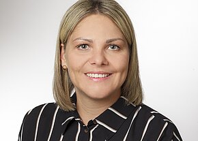 Porträtfoto einer lächelnden erwachsenen Frau mit schulterlangen blonden Haaren, vor neutralem Hintergrund. Sie trägt eine schwarz-weiße, vertikal gestreifte Bluse und kleine goldene Ohrringe, blickt direkt in die Kamera.