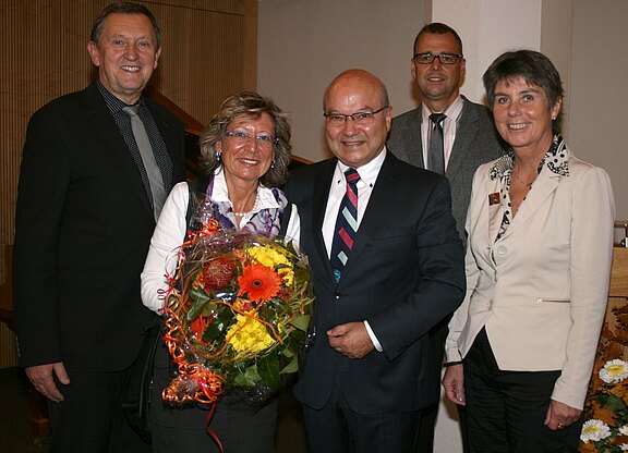 Landrat Herrmann Hübner, Oberbürgermeisterin Brigitte Merk-Erbe und Geschäftsführer Dr. Joachim Haun dankten Professor Dr. Dieter Hausmann für sein herausragendes Engagement. Fünf Erwachsene in festlicher Kleidung posieren lächelnd in einem Innenraum; die Frau links der Mitte hält einen großen Blumenstrauß mit orange-gelben Blüten. Die Szene wirkt wie eine Gratulation oder Ehrung.