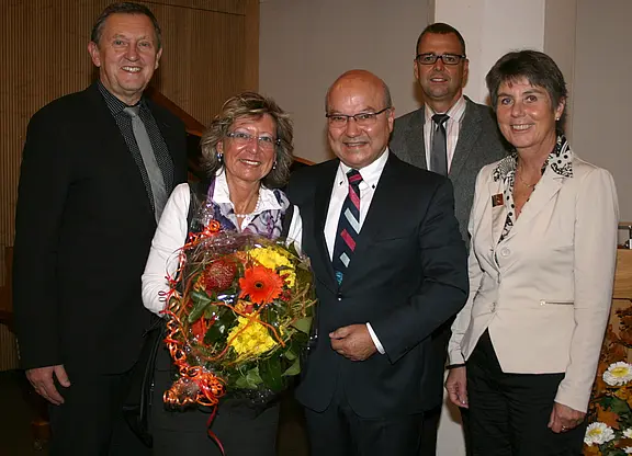 Fünf Erwachsene in festlicher Kleidung posieren lächelnd in einem Innenraum; die Frau links der Mitte hält einen großen Blumenstrauß mit orange-gelben Blüten. Die Szene wirkt wie eine Gratulation oder Ehrung.