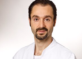 Porträt eines Mannes mit dunklen, zurückgekämmten Haaren und kurzem Bart. Er blickt ruhig in die Kamera und trägt einen weißen Kittel. Heller, neutraler Hintergrund.