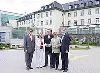 Fünf Personen stehen lächelnd vor einem Gebäude. Die Gruppe setzt sich aus Männern und einer Frau zusammen, die in formeller Kleidung gekleidet sind. Sie halten sich an den Händen und scheinen eine Zusammenarbeit oder ein Ereignis zu feiern. Der Hintergrund zeigt ein modernes und traditionelles Gebäude.