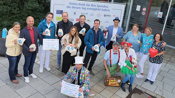 Spende für die Kinderklinik der Klinikum Bayreuth GmbH Gruppenfoto vor einem Gebäude: Krankenhausmitarbeitende und zwei Klinikclowns halten Spendenherzen und einen großen Scheck. Hinter ihnen ein Banner mit Dank an Spender. Stimmung fröhlich; Aktion offenbar Benefiz oder Spendenübergabe.
