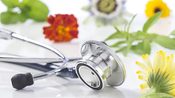 Ein silbernes Stethoskop liegt auf einer weißen Fläche, umgeben von bunten Heilblüten und Kräutern – Symbol für die Verbindung von Medizin und Naturheilkunde.