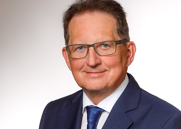 Porträt eines Mannes mit Brille, der in einem dunklen Anzug und einer gemusterten Krawatte gekleidet ist. Er hat short, brünette Haare und lächelt mild in die Kamera. Der Hintergrund ist hell und einfarbig.