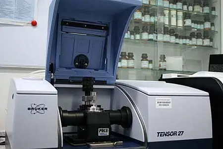 Das Bild zeigt ein wissenschaftliches Gerät, das als Fourier-Transformations-Infrarotspektrometer dient. Es hat eine kastenförmige Bauweise mit einem offenen Deckel und einem zentralen Halter für Proben. Im Hintergrund sind Regale mit Fläschchen sichtbar, die verschiedene chemische Substanzen enthalten.