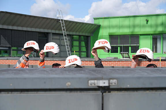 Mehrere Hände halten weiße Bauhelme mit roter Aufschrift über eine graue Kante. Es wirkt wie ein gemeinsamer Gruß von Bauarbeitern auf einer Baustelle.