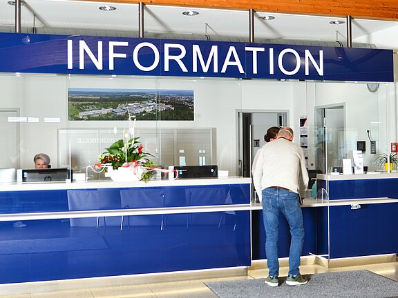 Ein Informationsschalter in einer Empfangshalle. Der Schalter ist blau mit dem Wort "INFORMATION" in großen Buchstaben. Eine Person steht am Schalter und spricht mit einer Mitarbeiterin, die hinter dem Schalter sitzt. Pflanzen dekorieren den Bereich.