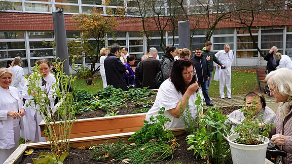 Mehrere Menschen in weißer Kleidung und ältere Personen versammeln sich im Innenhof um Hochbeete, betrachten und pflegen Kräuter und Gemüse. Es wirkt wie eine gemeinschaftliche Gartenaktion.