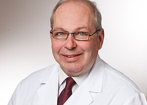 Dr. med. Wolfgang Withold Ein lächelnder Arzt trägt einen weißen Kittel und eine Krawatte. Er hat Brille und graue Haare. Der Hintergrund ist neutral und hell, was auf eine professionelle Atmosphäre hinweist.