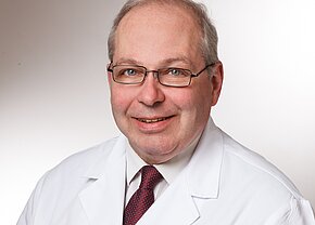 Ein lächelnder Arzt trägt einen weißen Kittel und eine Krawatte. Er hat Brille und graue Haare. Der Hintergrund ist neutral und hell, was auf eine professionelle Atmosphäre hinweist.