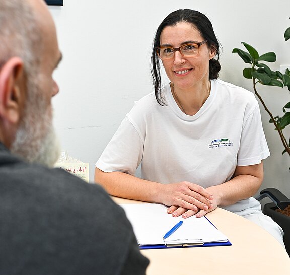 Ärztin im Gespräch mit Patient an kleinem runden Tisch. Sie trägt weiße Arbeitskleidung, lächelt und hört zu. Vor ihnen liegen Klemmbrett und Stift – Beratungssituation in einer Praxis.