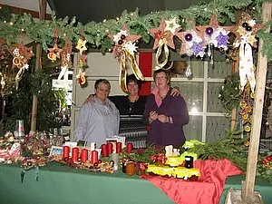 Drei Frauen stehen an einem festlich dekorierten Marktstand, der mit Weihnachtsschmuck und Kerzen geschmückt ist. Im Hintergrund sind Tannenzweige und Sterne zu sehen, die eine gemütliche, festliche Atmosphäre schaffen. Der Stand bietet eine Vielzahl von handgefertigten Weihnachtsartikeln an.