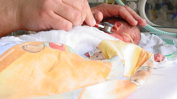 Ein Arzt oder eine Pflegekraft untersucht ein Frühgeborenes im Inkubator mit einem Stethoskop. Das Baby liegt unter einer Decke, hat eine Nasensonde und ist an Schläuche angeschlossen. Die Hand des Erwachsenen stützt behutsam den Kopf.