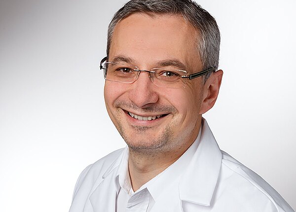 Porträt einer lächelnden Person mit Brille und kurzem Haar, die einen weißen Kittel trägt; neutraler, heller Hintergrund.