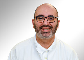 Porträt eines lächelnden Mannes mit Brille und kurzem Bart, kahlköpfig, in weißem Oberteil vor hellem, neutralem Hintergrund.