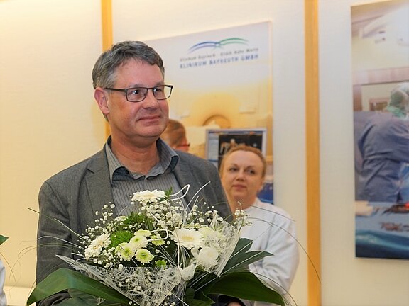 Ein Mann mit grauen Haaren und Brille hält einen Blumenstrauß in der Hand. Er trägt ein graues Sakko und lächelt leicht. Im Hintergrund sind unscharfe Bilder zu sehen, die eine Krankenhausumgebung darstellen. Eine Frau in einem weißen Kittel schaut interessiert.
