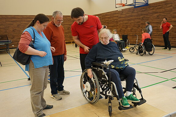 Übungsstunde in der Sporthalle: Ein Trainer erklärt einer Frau im Rollstuhl eine Fahrtechnik über eine Holzrampe, während zwei Teilnehmende aufmerksam zusehen. Im Hintergrund üben weitere Personen in Rollstühlen.