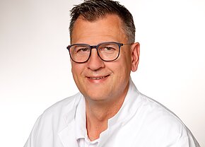 Dr. med. Markus Doreth Ein lächelnder Mann mit Brille und kurzen Haaren trägt einen weißen Kittel. Er hat einen freundlichen Ausdruck und steht vor einem hellen Hintergrund.