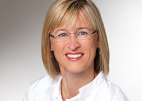 Porträt einer lächelnden Frau mit blondem, schulterlangem Haar und Brille. Sie trägt ein weißes Oberteil und blickt direkt in die Kamera vor einem neutralen, hellen Hintergrund.