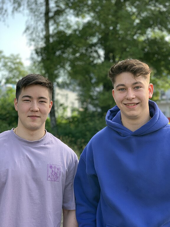 Zwei junge Personen stehen nebeneinander im Freien und schauen in die Kamera. Links ein Mann mit kurzem dunklem Haar in hellviolettem T‑Shirt, rechts ein lächelnder Mann im blauen Hoodie. Unscharfer, grüner Hintergrund.