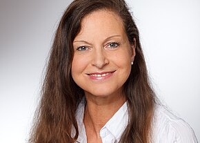 Sylvia Nützel Porträtfoto: Eine lächelnde erwachsene Frau mit langen braunen Haaren und Perlenohrringen blickt in die Kamera. Sie trägt eine weiße Bluse. Neutraler, heller Hintergrund.