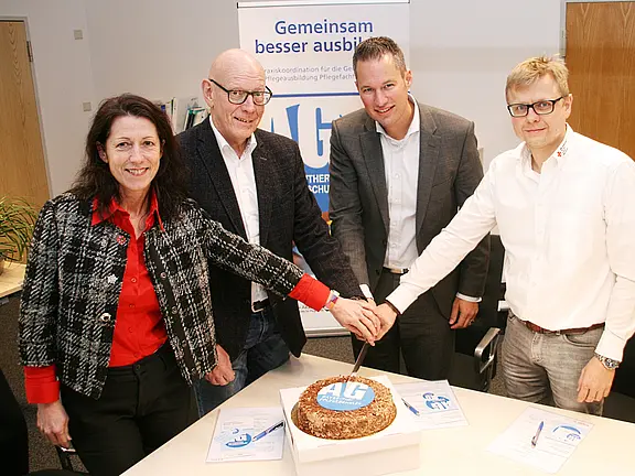 Vier Personen stehen um einen Tisch und schneiden gemeinsam eine Torte an, die mit einem Logo verziert ist. Im Hintergrund ist ein Banner mit dem Schriftzug „Gemeinsam besser ausbilden“ sichtbar. Die Atmosphäre wirkt feierlich und kooperativ.