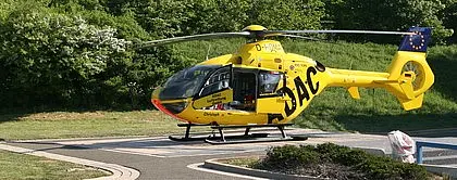 Gelber ADAC-Rettungshubschrauber steht auf einem Helipad im Freien; Seitentür geöffnet, medizinische Ausrüstung sichtbar. Grünflächen im Hintergrund.