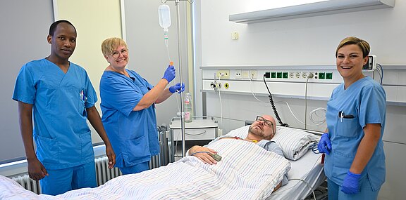 Krankenhauszimmer: Ein Patient liegt entspannt im Bett und erhält eine Infusion. Drei Pflegekräfte in blauen Kitteln stehen daneben, lächeln und überwachen die Behandlung.