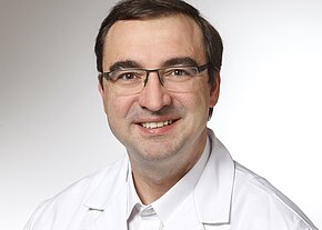 Dr. med. Kurt Wörner, MHBA Porträt eines lächelnden erwachsenen Mannes mit Brille in weißem Kittel vor neutralem Hintergrund, Kopf und Oberkörper frontal.