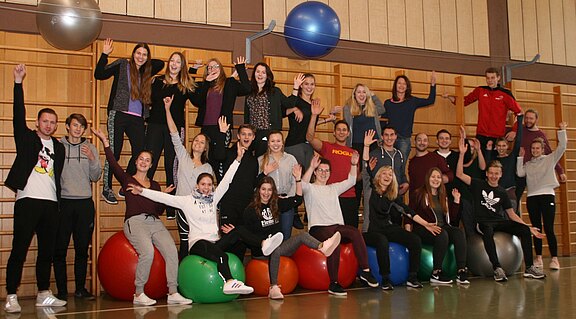 Große Gruppe junger Menschen in einer Sporthalle. Viele sitzen auf bunten Gymnastikbällen und werfen jubelnd die Arme hoch. Die Stimmung ist fröhlich und energiegeladen, wie bei einem Fitnesskurs oder Teamevent.