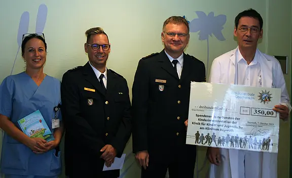 Zwei Polizisten, eine Pflegekraft und ein Arzt präsentieren einen großen Spenden-Scheck. Betrag: 350 Euro für die Kinder- und Jugendklinik. Die Gruppe steht zusammen im Krankenhaus und lächelt in die Kamera.