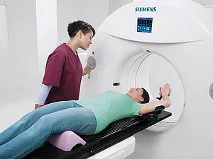 Eine medizinische Fachkraft steht neben einem Patienten, der auf einer Liege liegt und sich in einer MRT-Maschine befindet. Der Patient trägt ein grünes T-Shirt und hat die Arme seitlich angelegt. Die Fachkraft spricht mit dem Patienten, während das Gerät bereit ist, zu arbeiten.