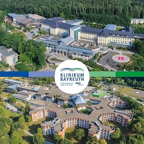 Luftaufnahme des Klinikum Bayreuth, die zwei Gebäudeansichten in einem grünen Umfeld zeigt. Im oberen Bereich ist ein modernes Gebäude zu sehen, während im unteren Bereich eine weitere Institution des Klinikums abgebildet ist, darunter das Logo „Klinikum Bayreuth“.