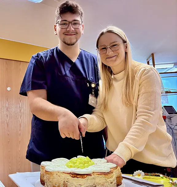 Zwei lächelnde Personen schneiden gemeinsam eine große, runde Torte an. Der links Stehende trägt dunkelblaue Arbeitskleidung, die rechts Stehende einen hellen Pullover. Auf der Torte liegen helle Obststücke. Fokus: gemeinsamer Anschnitt bei einer Feier.