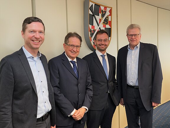 Vier Männer stehen nebeneinander, freundlich lächelnd, in einem Innenraum. Drei tragen Anzüge mit Krawatte, einer Sakko mit offenem Hemd. Hinter ihnen hängt ein Wappen an der Wand. Gruppenfoto in einem formellen, beruflichen Kontext.