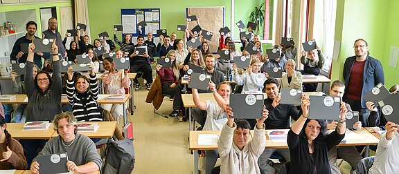 Eine große Gruppe von Menschen sitzt in einem Klassenzimmer und hält Bücher oder Ordner mit dem Aufkleber "BFS" in der Höhe. Sie lächeln und zeigen Freude, während einige in die Kamera schauen. Die Wände sind grün gestrichen, und es gibt Tische mit Unterlagen vor ihnen.
