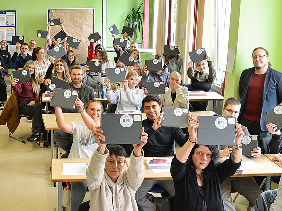 Eine große Gruppe von Menschen sitzt in einem Klassenzimmer und hält Bücher oder Ordner mit dem Aufkleber "BFS" in der Höhe. Sie lächeln und zeigen Freude, während einige in die Kamera schauen. Die Wände sind grün gestrichen, und es gibt Tische mit Unterlagen vor ihnen.