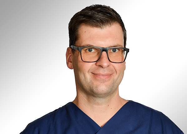 Porträt eines Mannes mit dunkler Brille und kurzem Haar, in dunkelblauer OP‑Kleidung, leicht lächelnd vor neutral-hellem Hintergrund.