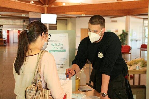Eine Person mit Brille und Maske steht vor einem Tisch, an dem ein Mann in einem schwarzen Shirt ebenfalls eine Maske trägt. Auf dem Tisch steht ein Schild mit der Aufschrift "Wäscheservice", das Informationen zu Öffnungszeiten enthält. Der Mann erklärt etwas und zeigt auf ein Formular.
