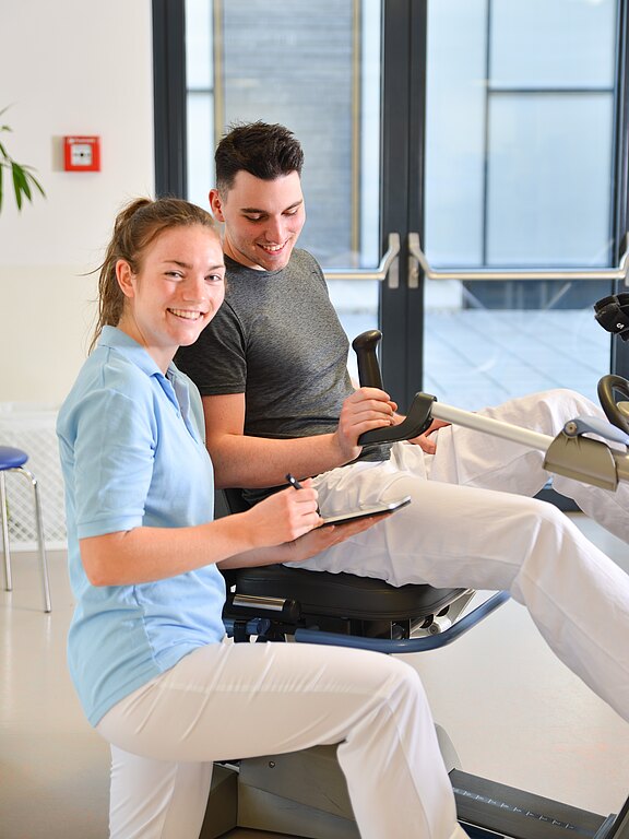 Therapeutin betreut einen Mann auf einem sitzenden Ergometer. Sie hält ein Klemmbrett und notiert Trainingsdaten, während er tritt. Beide wirken freundlich; die Szene deutet auf Reha oder physiotherapeutisches Training hin.