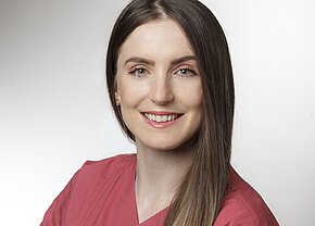 Xhesika Blloshmi Porträt einer lächelnden Frau mit langen, glatten Haaren in einem rosa Oberteil. Der Hintergrund ist hell und neutral. Die Frau hat grüne Augen und trägt kleine Ohrringe. Sie strahlt Freundlichkeit und Professionalität aus.