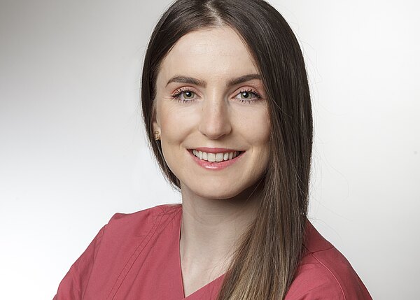 Porträt einer lächelnden Frau mit langen, glatten Haaren in einem rosa Oberteil. Der Hintergrund ist hell und neutral. Die Frau hat grüne Augen und trägt kleine Ohrringe. Sie strahlt Freundlichkeit und Professionalität aus.