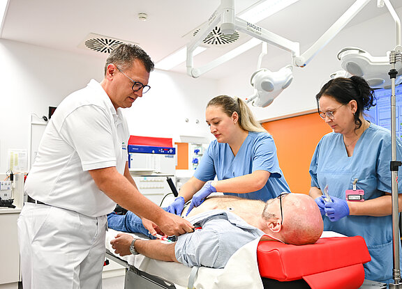 Drei medizinische Fachkräfte führen eine Untersuchung an einem Patienten durch, der auf einer Liege liegt. Der Patient ist teilweise entblößt, und einer der Helfer befestigt Elektroden an seiner Brust. Die Umgebung wirkt modern und klinisch.