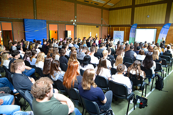 In einem großen Raum sitzen zahlreiche Menschen auf Stühlen und hören einer Präsentation zu. Auf einer Leinwand im Vordergrund sind Grafiken oder Texte sichtbar. Die Atmosphäre ist aufgeschlossen und konzentriert, während die Teilnehmer aufmerksam dem Vortrag folgen.