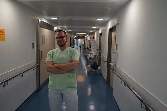Medizinische Fachkraft in grüner Arbeitskleidung steht mit verschränkten Armen in einem hellen Krankenhausflur. Im Hintergrund Türen und medizinische Wagen; klarer, sauberer Gang.