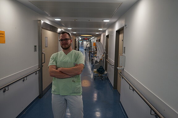 Medizinische Fachkraft in grüner Arbeitskleidung steht mit verschränkten Armen in einem hellen Krankenhausflur. Im Hintergrund Türen und medizinische Wagen; klarer, sauberer Gang.