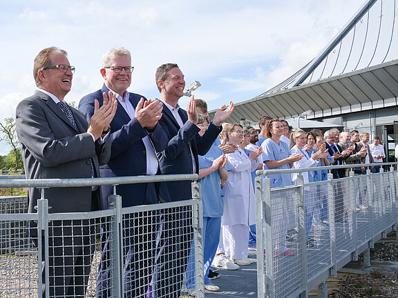 Eine Gruppe von Menschen, teils in Anzügen, teils in Klinikbekleidung, steht im Freien hinter einem Geländer und applaudiert vor einem modernen Gebäudeteil unter blauem Himmel.