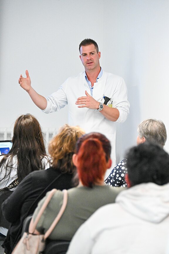 Ein Mann im weißen Kittel spricht vor einer sitzenden Gruppe und gestikuliert, als halte er einen medizinischen Vortrag oder eine Schulung in einem hellen Raum.