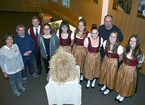 Gruppenfoto in einem Saal: Mehrere junge Frauen in braun-roten Trachten und weitere Personen stehen um einen Tisch. Darauf eine große, kunstvoll gebundene Erntekrone aus Stroh, zentraler Blickfang.